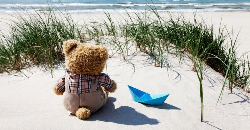 Ein Teddy sitzt mit einem blauen Papierschiff am Strand